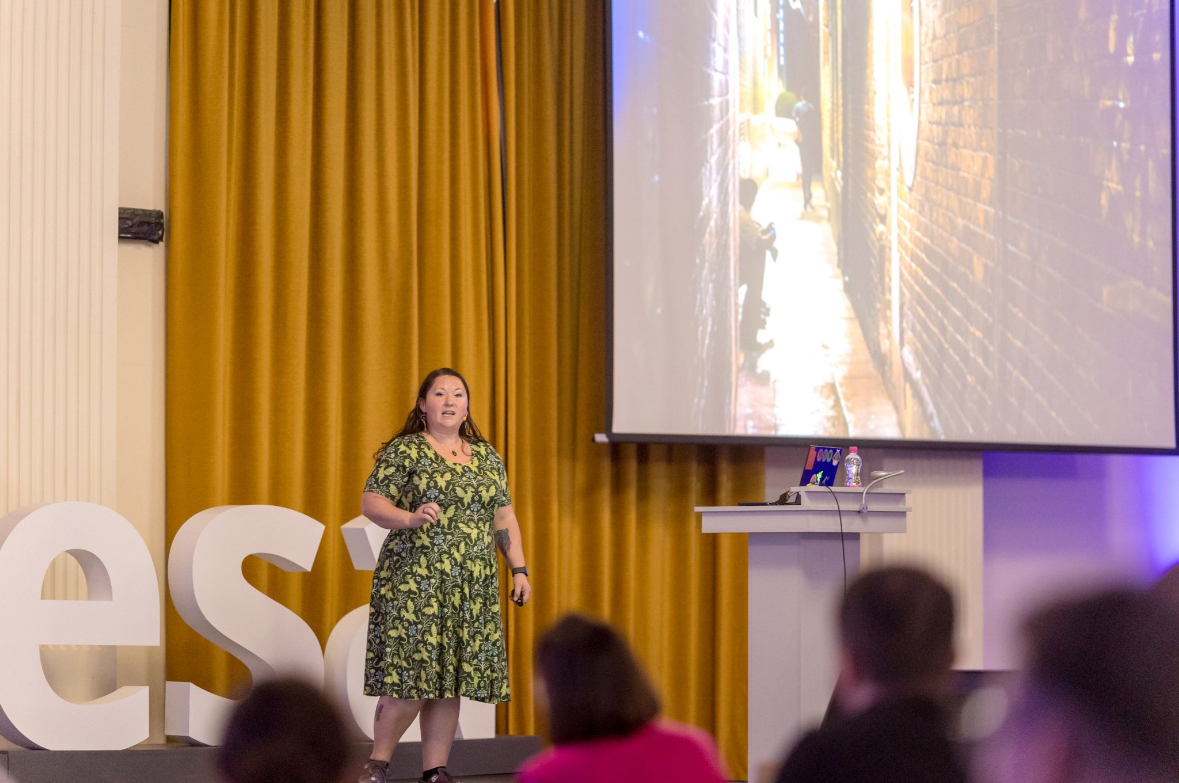 Rachel presenting on a conference stage in a green leaf-patterned dress, a projection screen and audience beside her