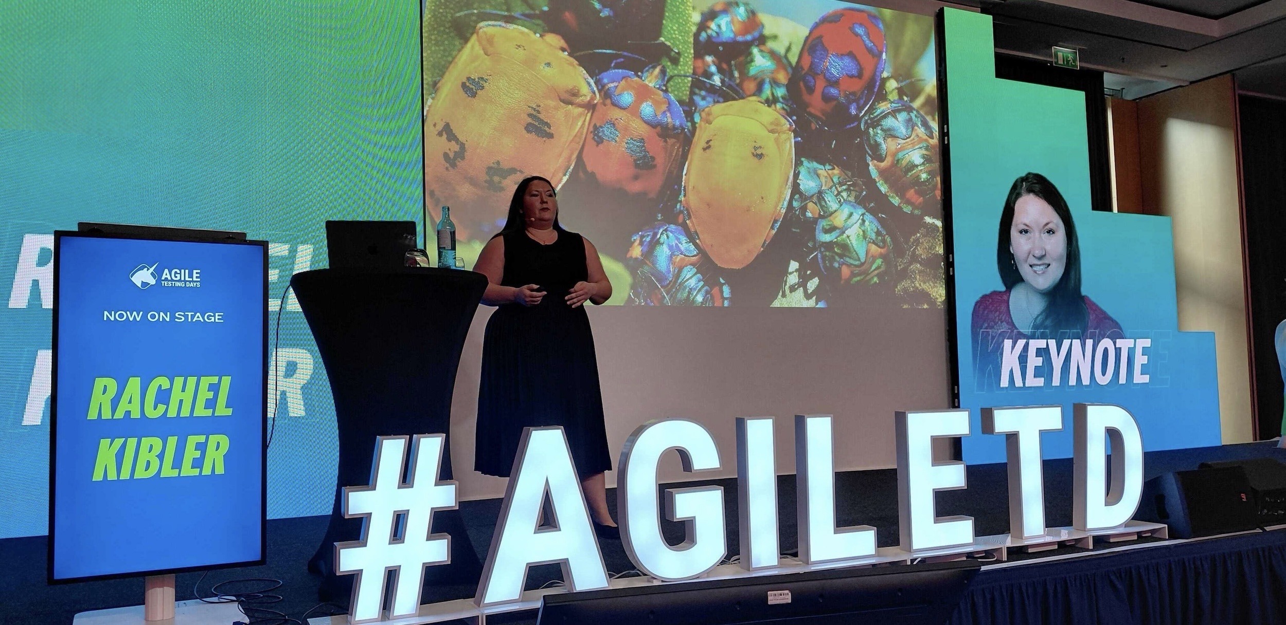 Rachel Joi Kibler speaking on stage at Agile Testing Days, standing behind giant illuminated #AGILETD letters with a 'Now on stage: Rachel Kibler' screen beside her