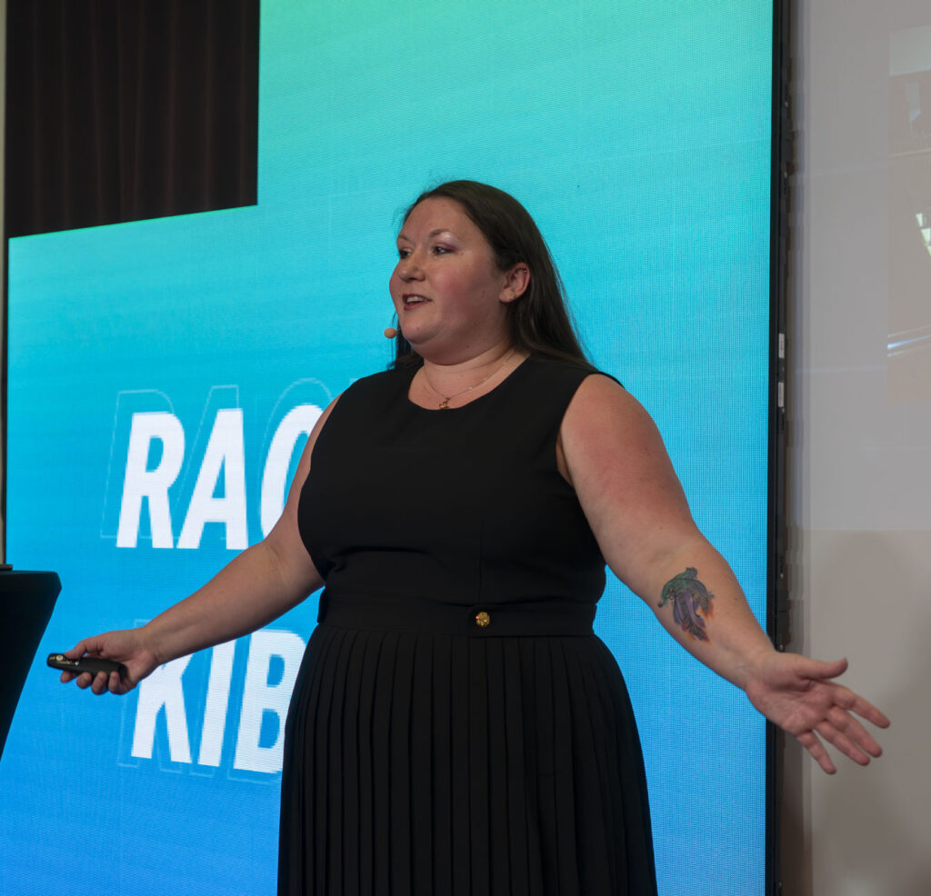 Rachel speaking on stage in a black dress, a teal LED wall displaying her name behind her