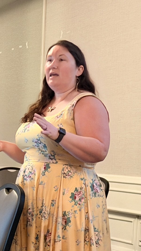 Rachel speaking in a conference room, gesturing, wearing a yellow floral sundress