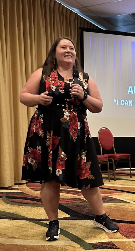 Rachel holding a microphone as she speaks, wearing a black dress with a red floral pattern