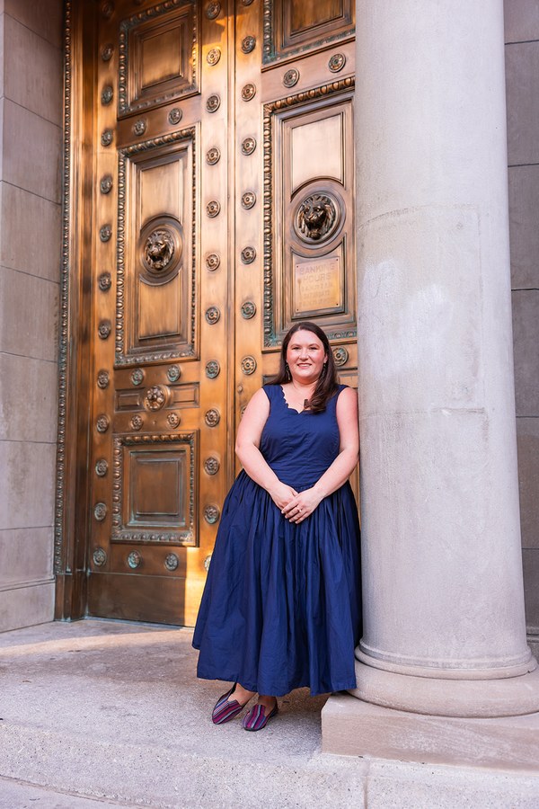 Rachel standing beside a stone column in a navy dress, tall bronze doors behind her