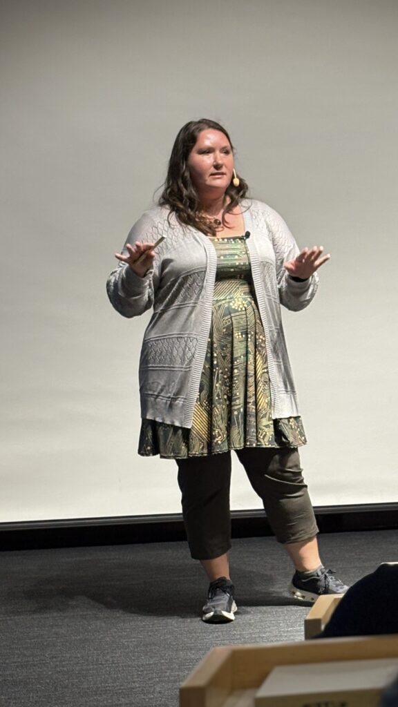 Rachel speaking on stage, gesturing with both hands, wearing a grey cardigan over a green patterned dress